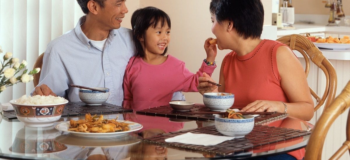 An Asian family, an adult male and female are seated around a table eating a meal with a young female standing in between the adults.